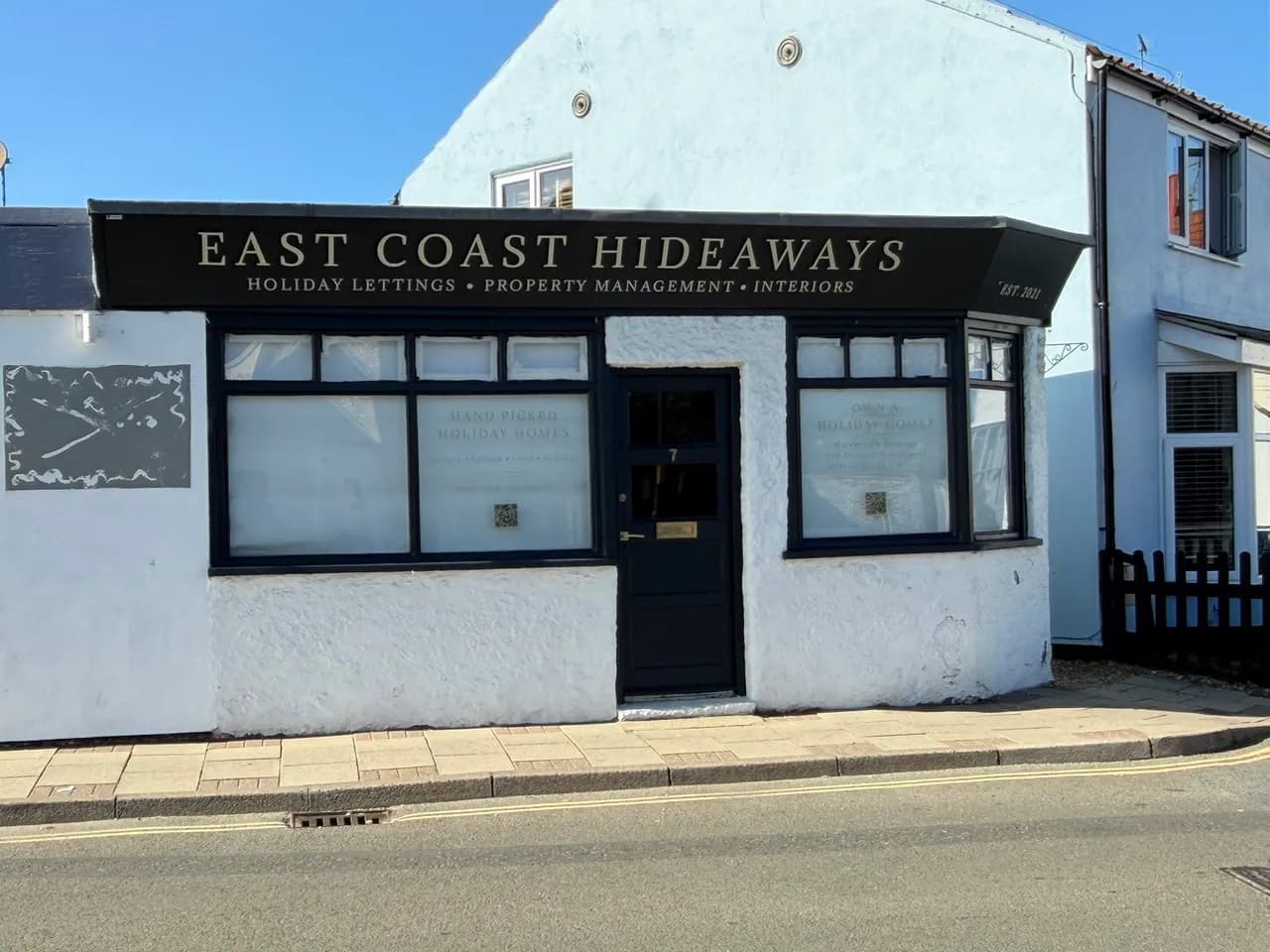 A light blue and white building with a black awning reading 'EAST COAST HIDEAWAYS' over two windows and a dark blue door.