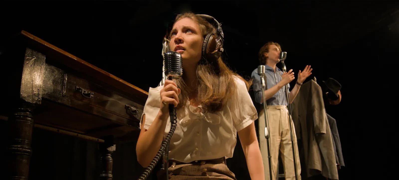 A woman with headphones and a vintage microphone looks up with a man singing in the background.