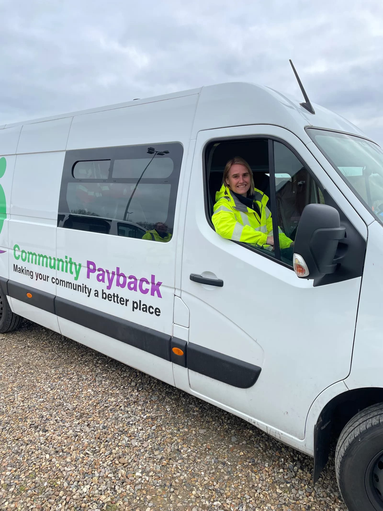 A smiling woman in a bright yellow safety jacket leans out of the driver's side window of a white van that says 'Community Payback' on its side.