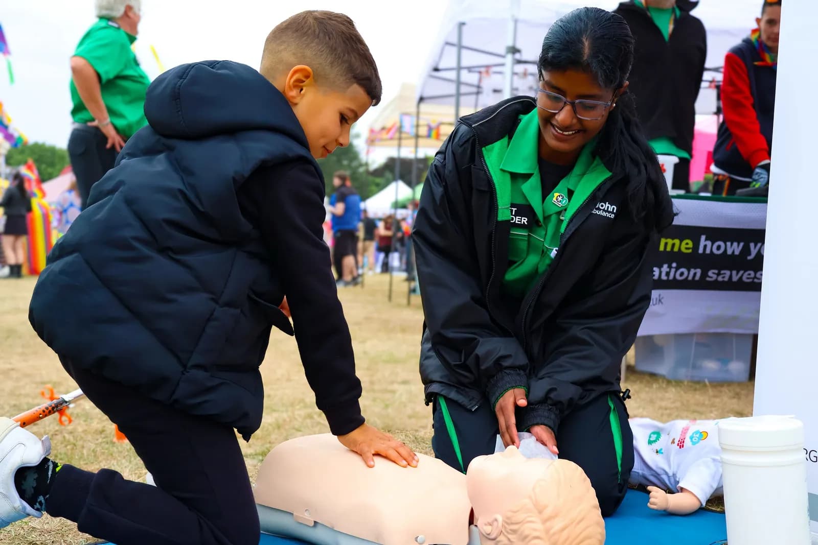 A smiling woman in a green and black uniform demonstrates CPR on a dummy, while a young boy observes, leaning over the dummy.