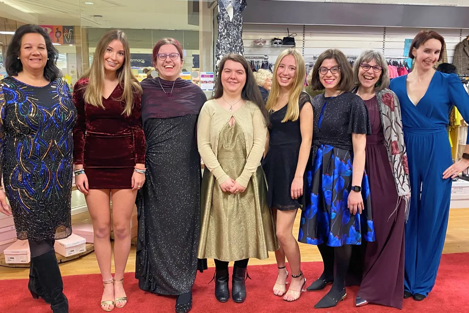 Eight women are standing side by side on a red carpet, all smiling and wearing different formal attire.